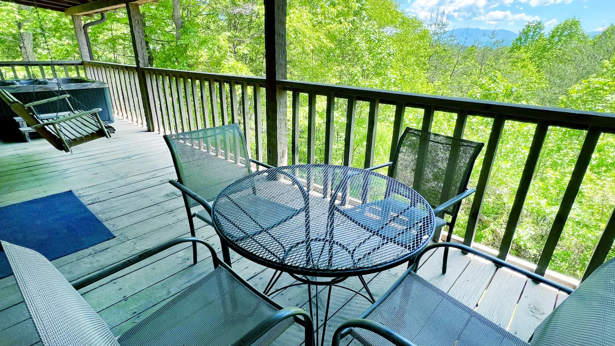The image shows the exterior of a cozy cabin in the Smoky Mountains, featuring a private deck with a table and chairs perfect for gathering, a bubbling hot tub overlooking lush greenery, and open views ideal for sunrise watching or stargazing at night.