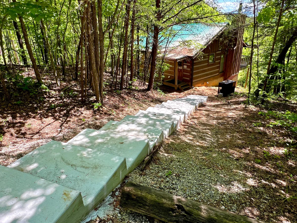 This exterior image features the cabin nestled among trees with a staircase leading to the entrance, and a tranquil lake view in the background. The natural setting suggests opportunities for relaxation after a day of adventures and hints at the outdoor spaces available for gathering, grilling, or unwinding.