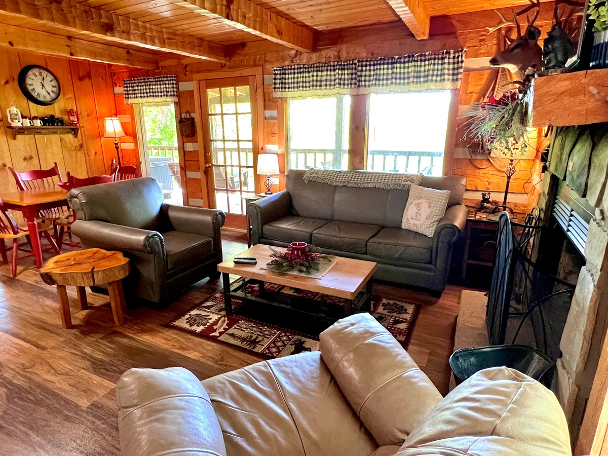 A broad view of the main living area reveals sofas arranged for socializing, a wooden dining table, a fireplace, and expansive windows that fill the space with natural light and offer glimpses of the outdoors. The rustic charm of wood accents is evident throughout, creating a seamless flow between relaxation and dining spaces.