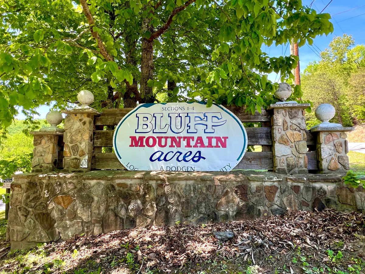 A welcoming exterior shot of the entrance to the Bluff Mountain Acres community, featuring prominent signage, stone pillars, and lush green foliage. The scene hints at both the natural beauty and the inviting atmosphere of the area.