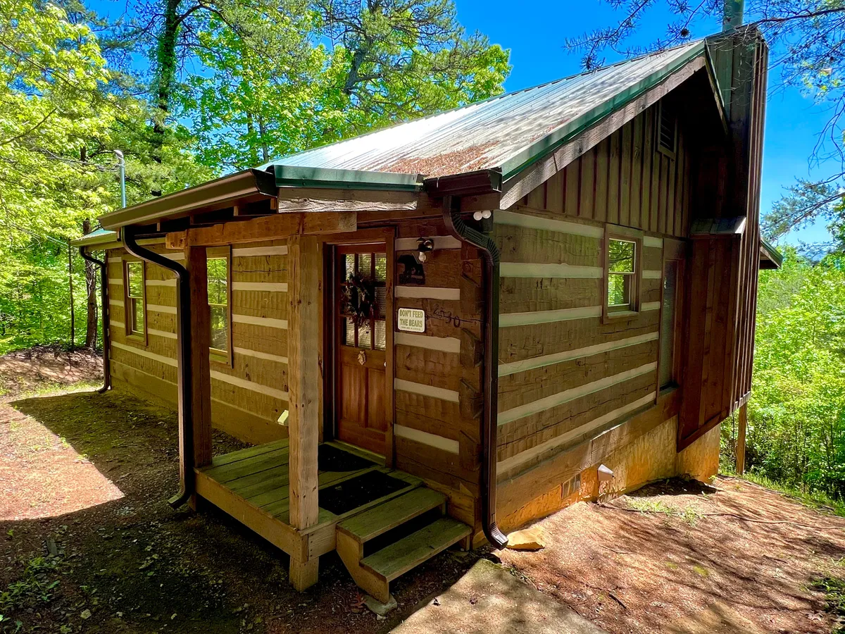 An appealing view of the cabin's front door, wooden steps, and rustic exterior surrounded by lush foliage. The inviting porch hints at homey comfort and a welcoming atmosphere for families arriving for their getaway.