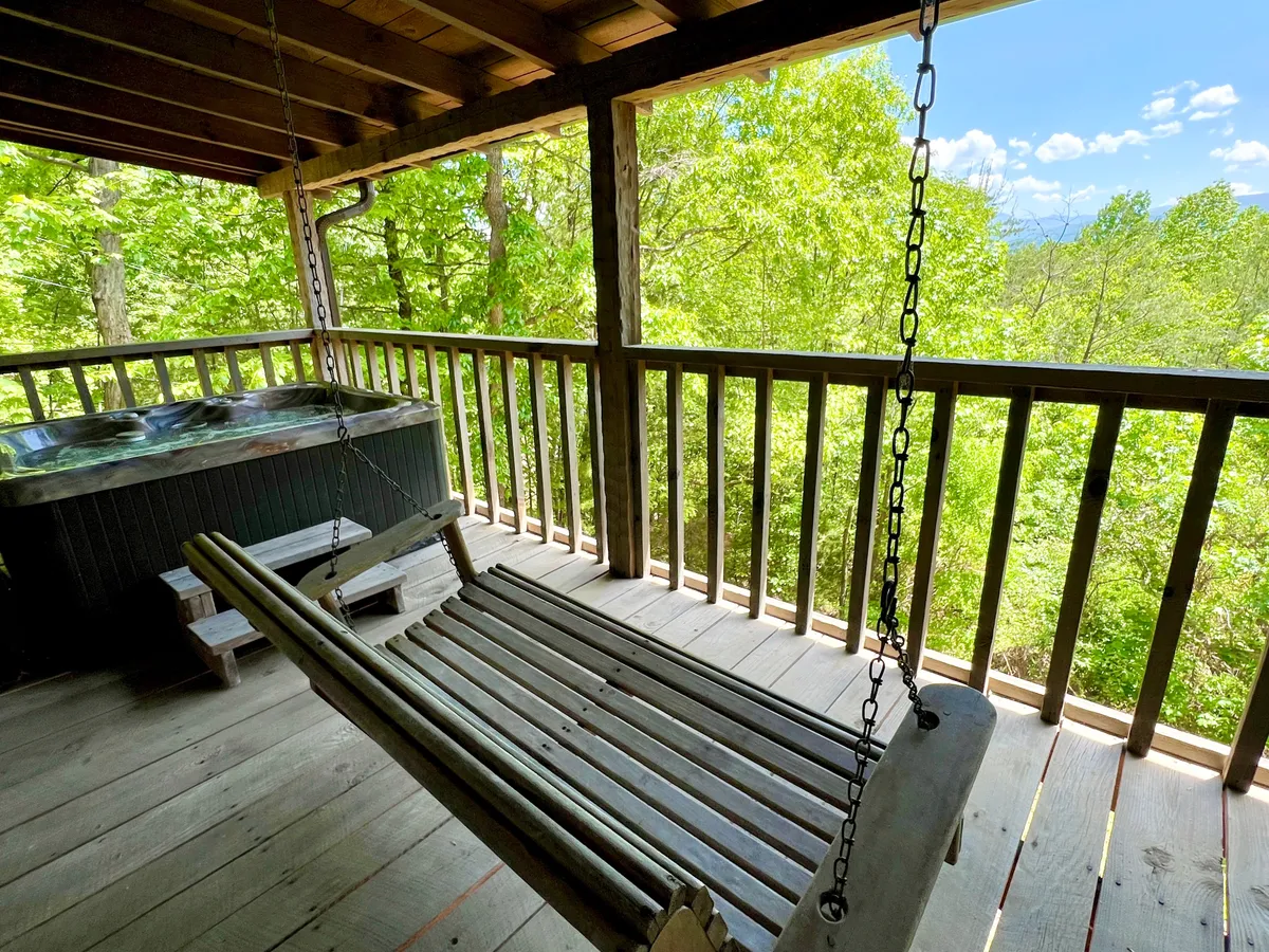 The scene presents a cozy swinging bench and a hot tub on the deck, set against a backdrop of lush greenery. The arrangement invites guests to alternate between gently swaying on the swing and soaking in the hot tub, all while immersed in the serenity of nature.