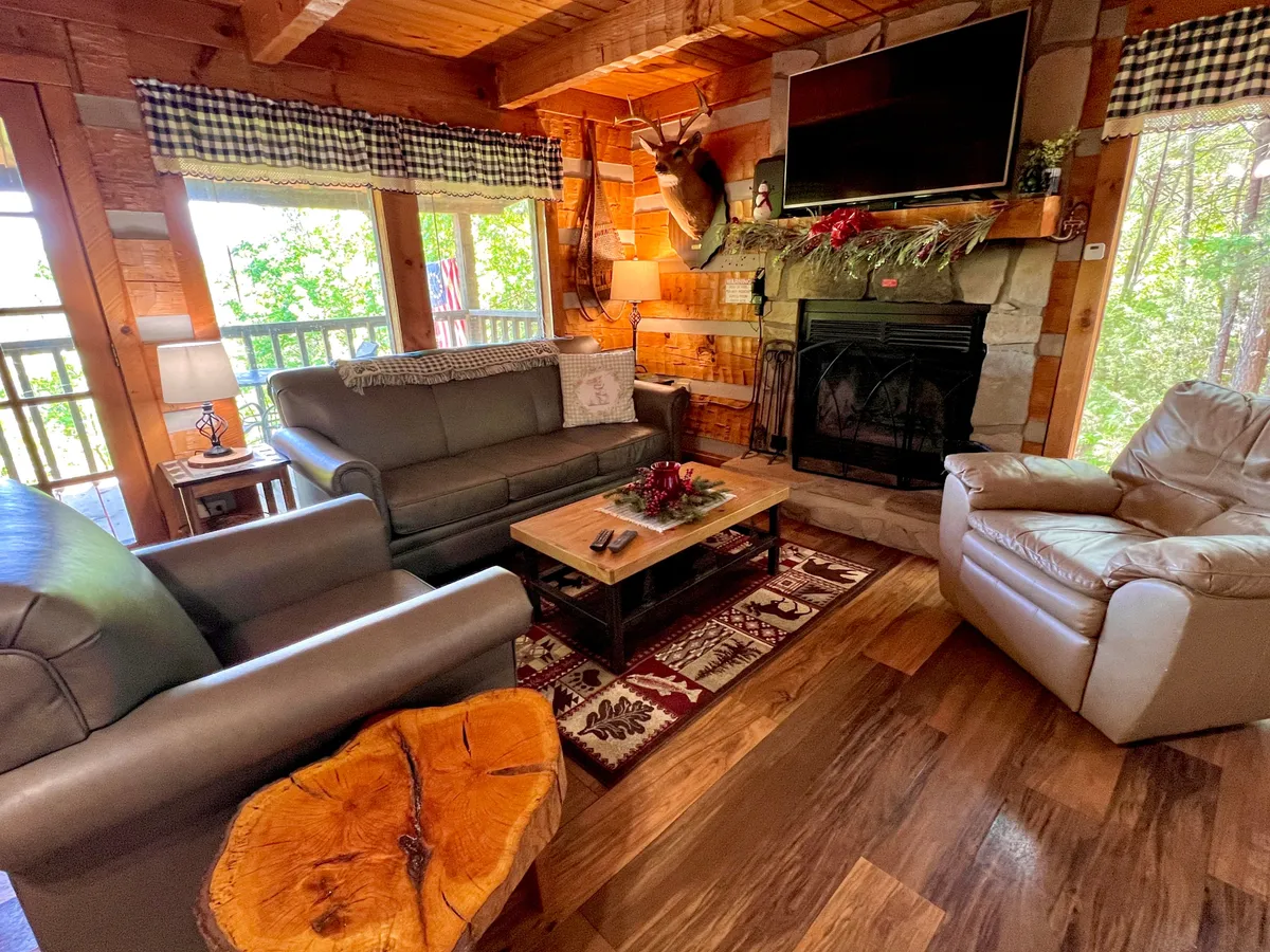 This image showcases an inviting living room with a stone-faced fireplace as the central focal point, surrounded by a comfortable sofa and armchair. The room features rustic wooden accents and décor that create a warm, welcoming atmosphere perfect for relaxing, reading, or gathering with family and friends.