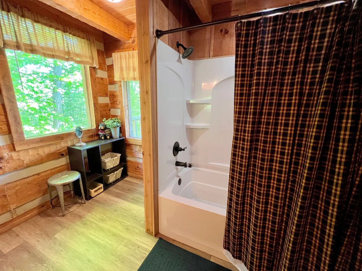 A full bathroom with wooden walls, a shower/tub combo with a curtain, natural light from windows, and a sleek black storage unit. The space looks freshly cleaned and inviting, with fluffy towels ready for guests’ comfort.