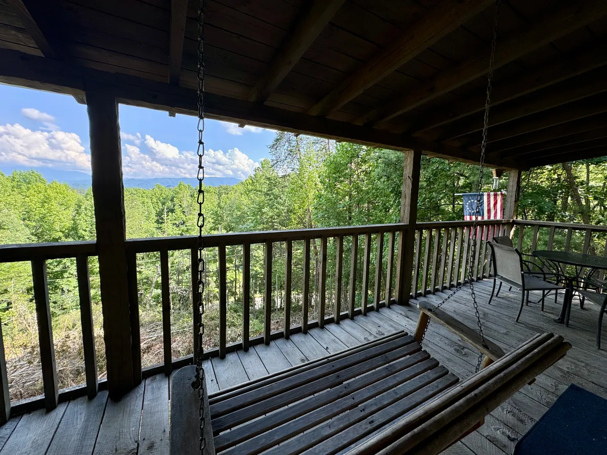 A wide view of the deck reveals a swinging bench positioned to take in the mountain views, with a wooden railing and greenery in the foreground. The American flag adds a touch of rustic Americana, and the open sky suggests opportunities for stargazing or enjoying sunsets.