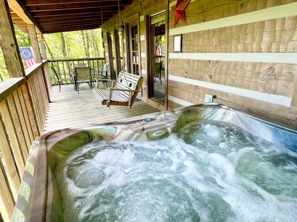 This image captures a tranquil exterior scene featuring a wooden deck equipped with a hot tub, comfortable seating area, and an American flag. The deck overlooks a lush tree line, offering a peaceful connection to nature and a private spot for relaxation after a day of adventure.