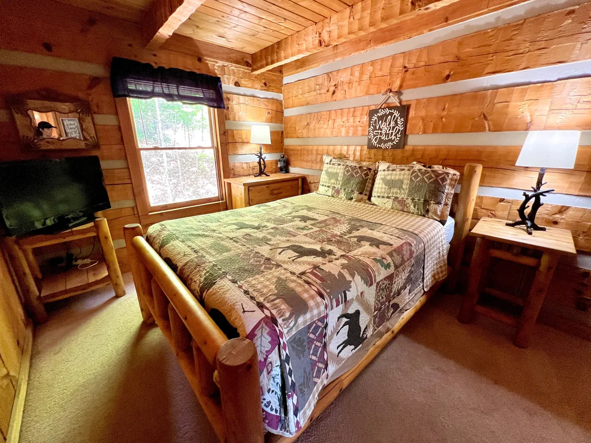 A warm and inviting bedroom featuring a cozy queen bed topped with a plush quilt, natural light streaming in from a window, and wooden furniture that offers both style and storage. Decorative wall art and soft linens create a restful, welcoming atmosphere.