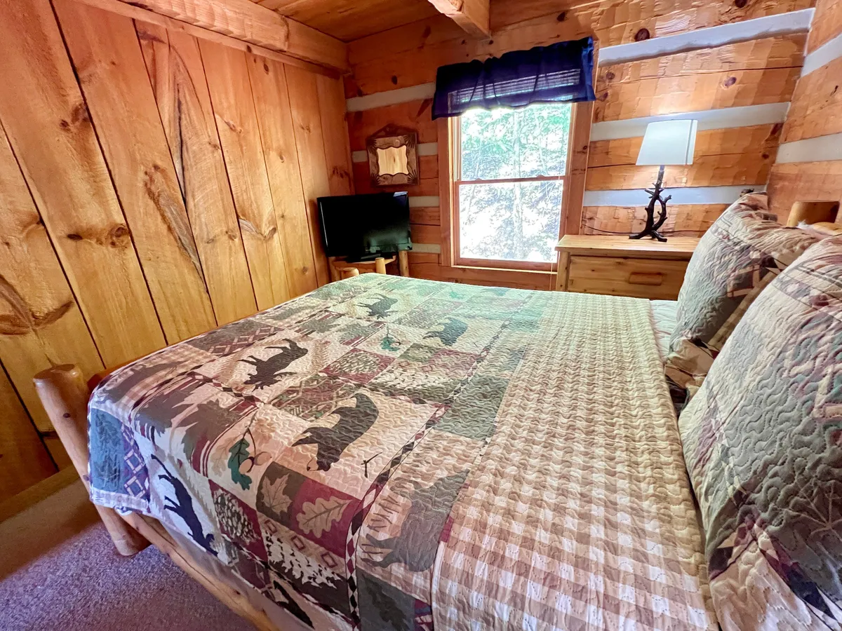 A tranquil bedroom with log cabin walls, a queen bed with a cozy quilt, and a nightstand lamp casting a soft glow. The window brings in natural light, enhancing the peaceful ambiance and highlighting the room's soothing décor.