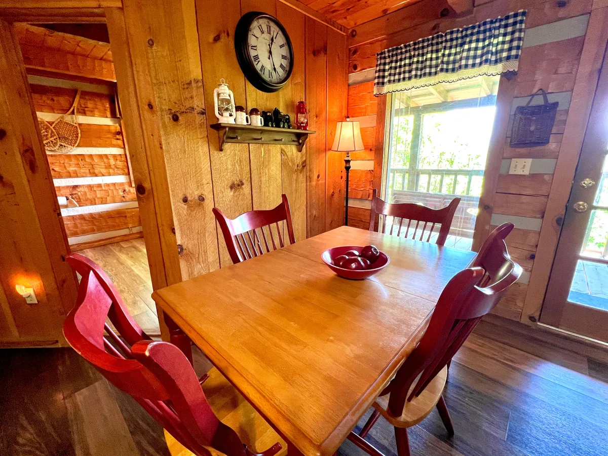 A bright dining area featuring a dining table for four with red chairs, a wall clock, a cozy lamp, and a window with a checkered curtain letting in natural light. The scene is set for family meals, game nights, or enjoying a morning coffee.