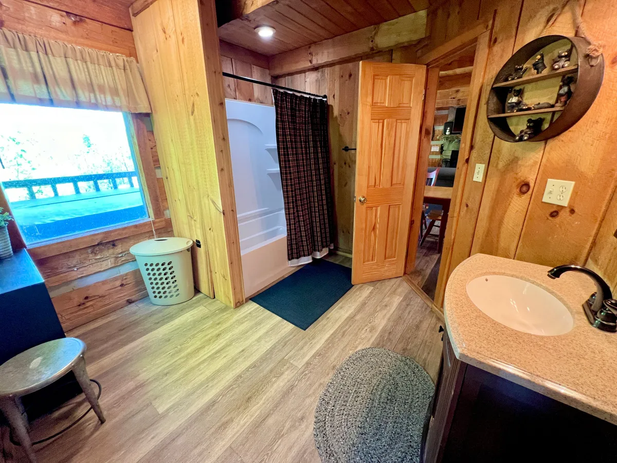 A view of the bathroom area with a vanity sink, shower area, wooden walls, a window, and a laundry basket in view. The layout suggests the presence of a washer/dryer, adding to the sense of convenience and practicality for guests.