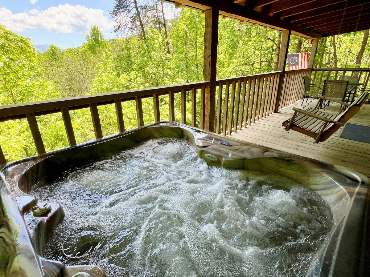 This image features a bubbling hot tub situated on a wooden deck, framed by the cabin's railing and surrounded by lush trees. The American flag adds a homey touch, while the open-air environment offers expansive, peaceful views of the Smoky Mountains. The scene is bathed in soft light, suggesting either morning or sunset, emphasizing relaxation and nature immersion.