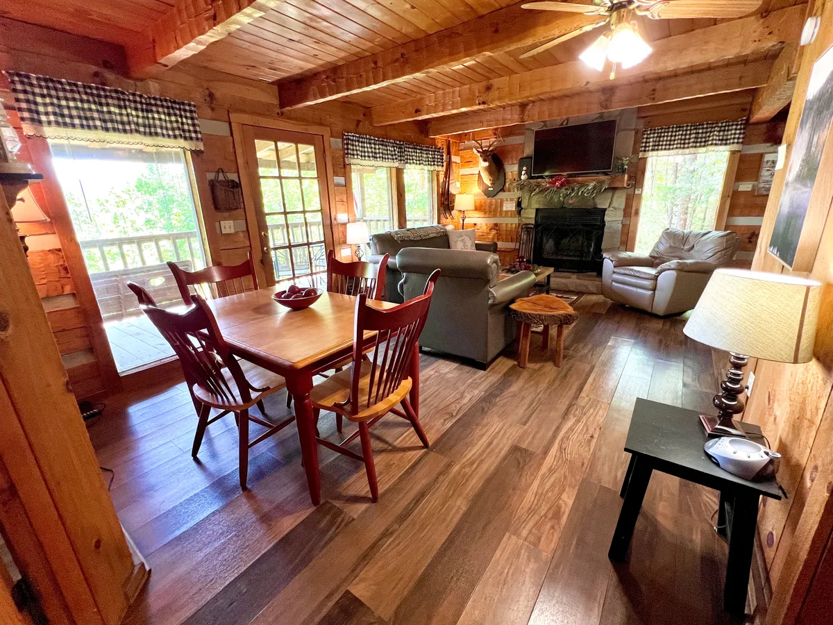 A spacious shared living area with a dining table, plush sofa, stone fireplace, and large windows that fill the space with natural light. The arrangement invites both relaxation and socializing, while the windows hint at the surrounding natural beauty.