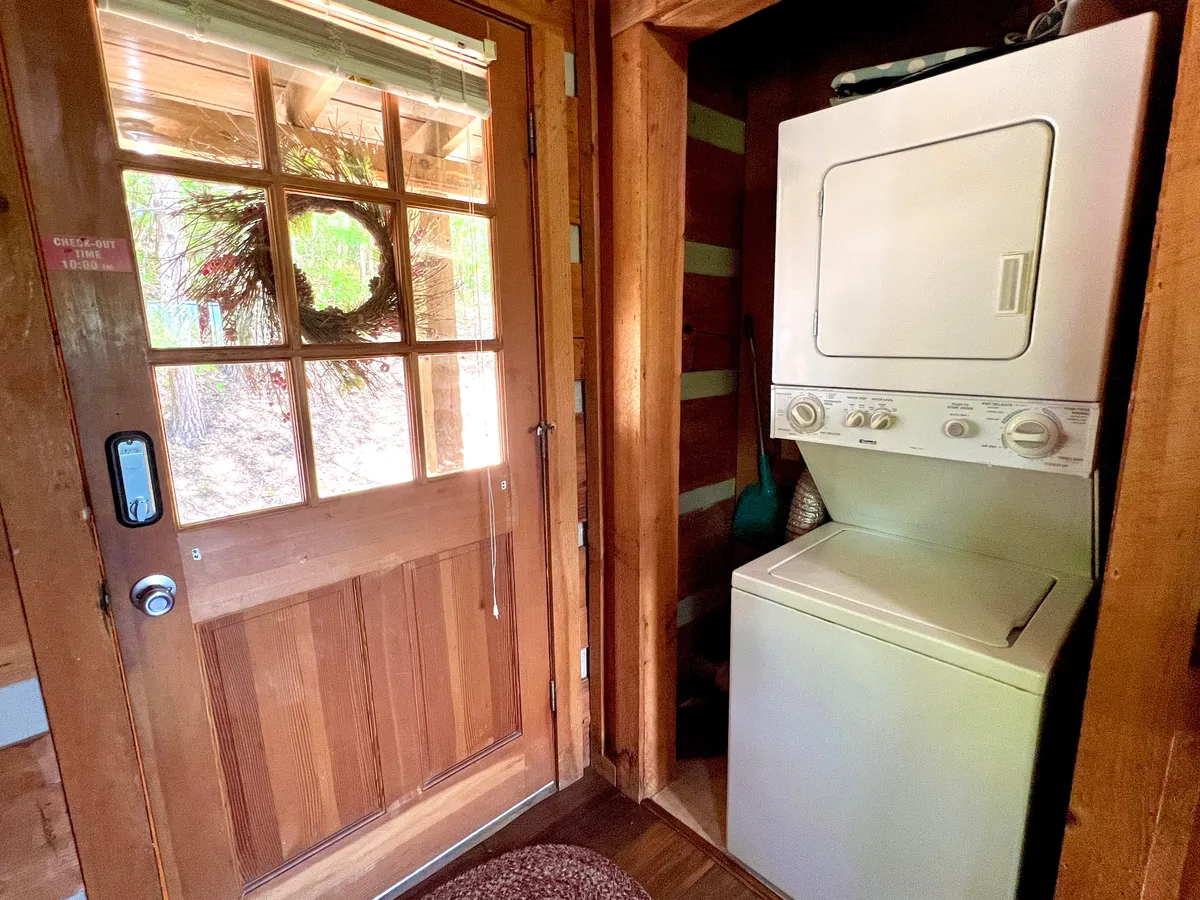 The entryway features a rustic wooden door, a decorative wreath, and a laundry machine tucked neatly to the side. The space is warmly lit and offers a welcoming first impression that encapsulates the ease of direct self check-in and immediate immersion into cabin comfort.